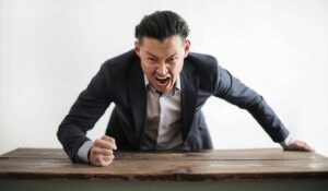 angry male employee in blue jacket shouting and pounding on wooden desk
