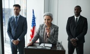 Older female executive speaking at podium flanked by two male security guards